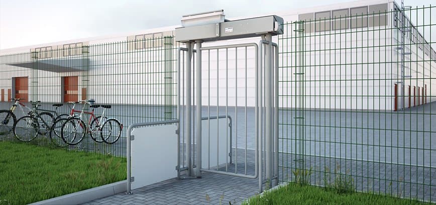 Magnetic MPG barrier-free swing gate installed in a mesh perimeter fence, with a cycle rack alongside and an industrial building beyond